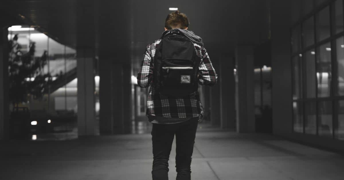 A young man in dark jeans, black and white plaid shirt, and a black backpack is seen from the back, walking alone.