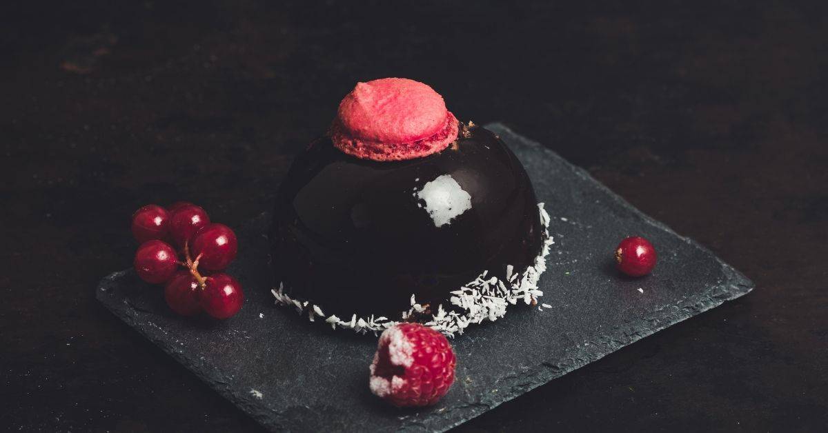 A dark mirror glazed dome cake topped with a red macaron, with sugar crystals around the bottom and flanked by red berries