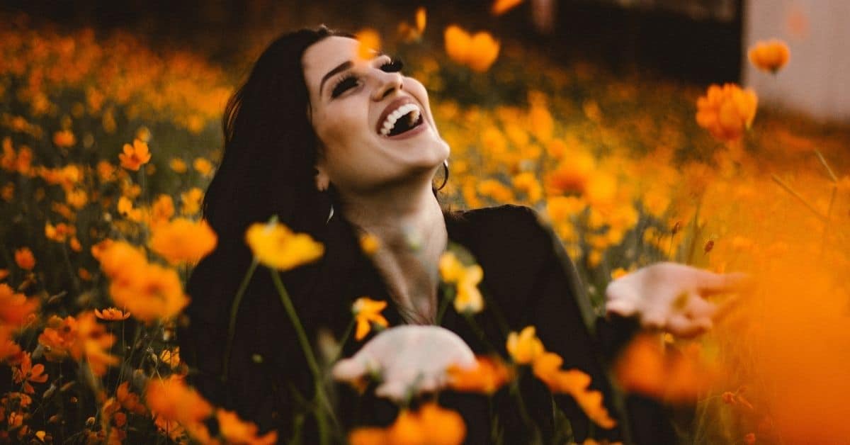 A dark haired woman in a black shirt laughing joyously in a field of yellow flowers.