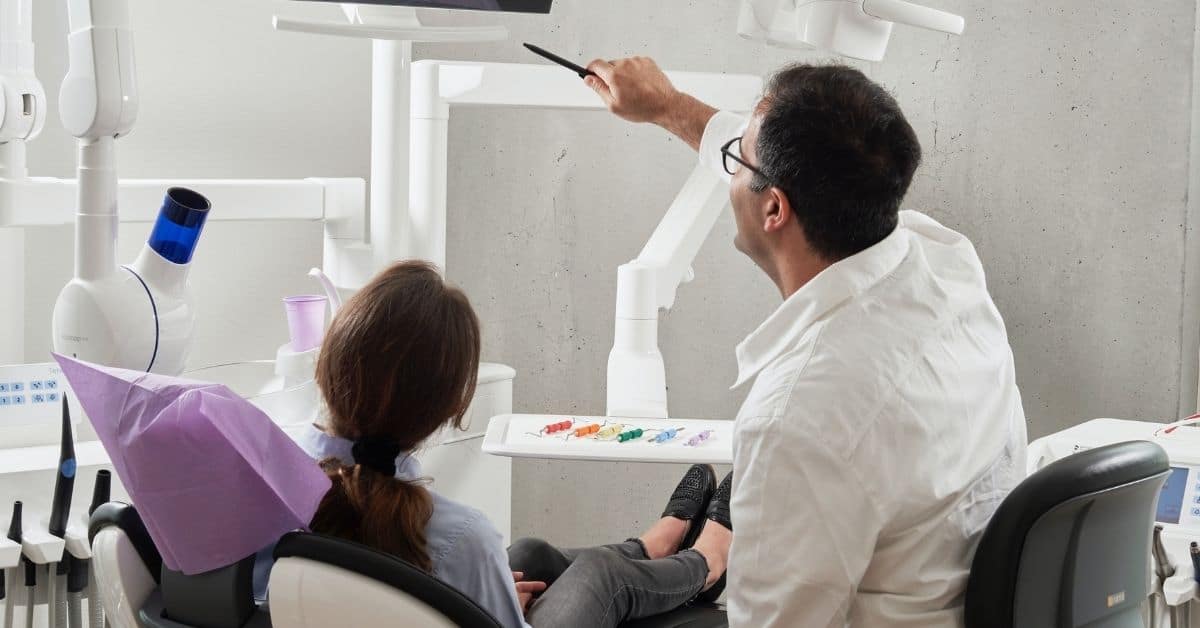 A male dentist in a white shirt pointing at an xray above a dark-haired woman in the patient chair.
